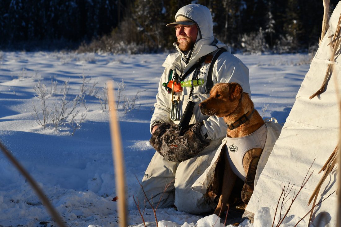 A hunter kneeling in snow beside a brown hunting dog in a white vest